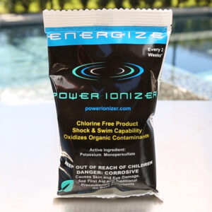 A packet of Energize Non-Chlorine Shock from Swimming Pool Discounters sits on a table near a pool, advertised as a chlorine-free maintenance product that oxidizes organic contaminants using potassium monopersulfate.