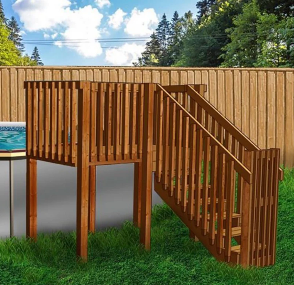 A wooden deck with railings and stairs stands on grass, next to a tall wooden fence, with trees and a cloudy blue sky in the background.