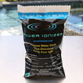 A bag of Awaken Pool Shock by Swimming Pool Discounters, featuring Potassium Monopersulfate for effective pool cleaning, stands upright on a poolside surface with a blurred swimming pool in the background. The packaging is black and blue with yellow and white text.