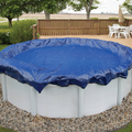 An above-ground pool with an SPD Standard Winter Cover is placed on a pebble border near a wooden deck with white patio furniture and an umbrella, surrounded by trees and grass in the background.