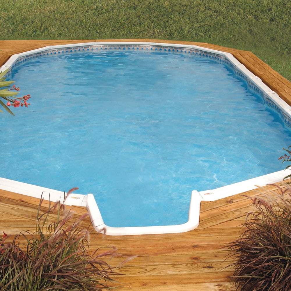 The Lomart 18' x 39' x 54" Oval Whispering Wind III Semi In-Ground Pool with In-Step Package sits on a wooden deck surrounded by green grass and ornamental plants, its galvanized steel walls holding clear blue water.
